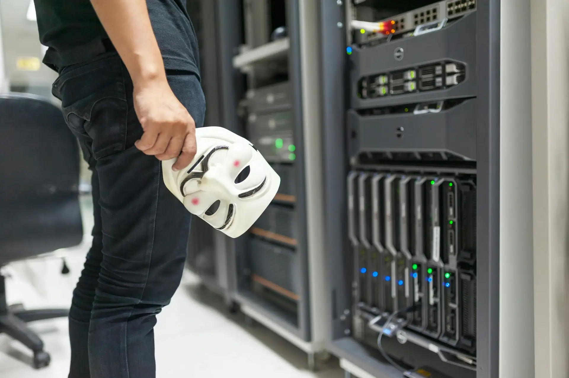 Home Person holding anonymous mask near servers, hinting at cybersecurity and hacking themes.